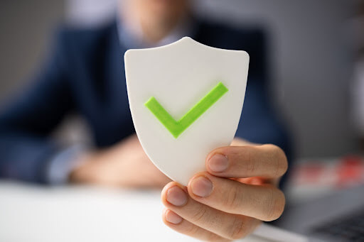 A hand holding a white shield with a green checkmark on it for business protection